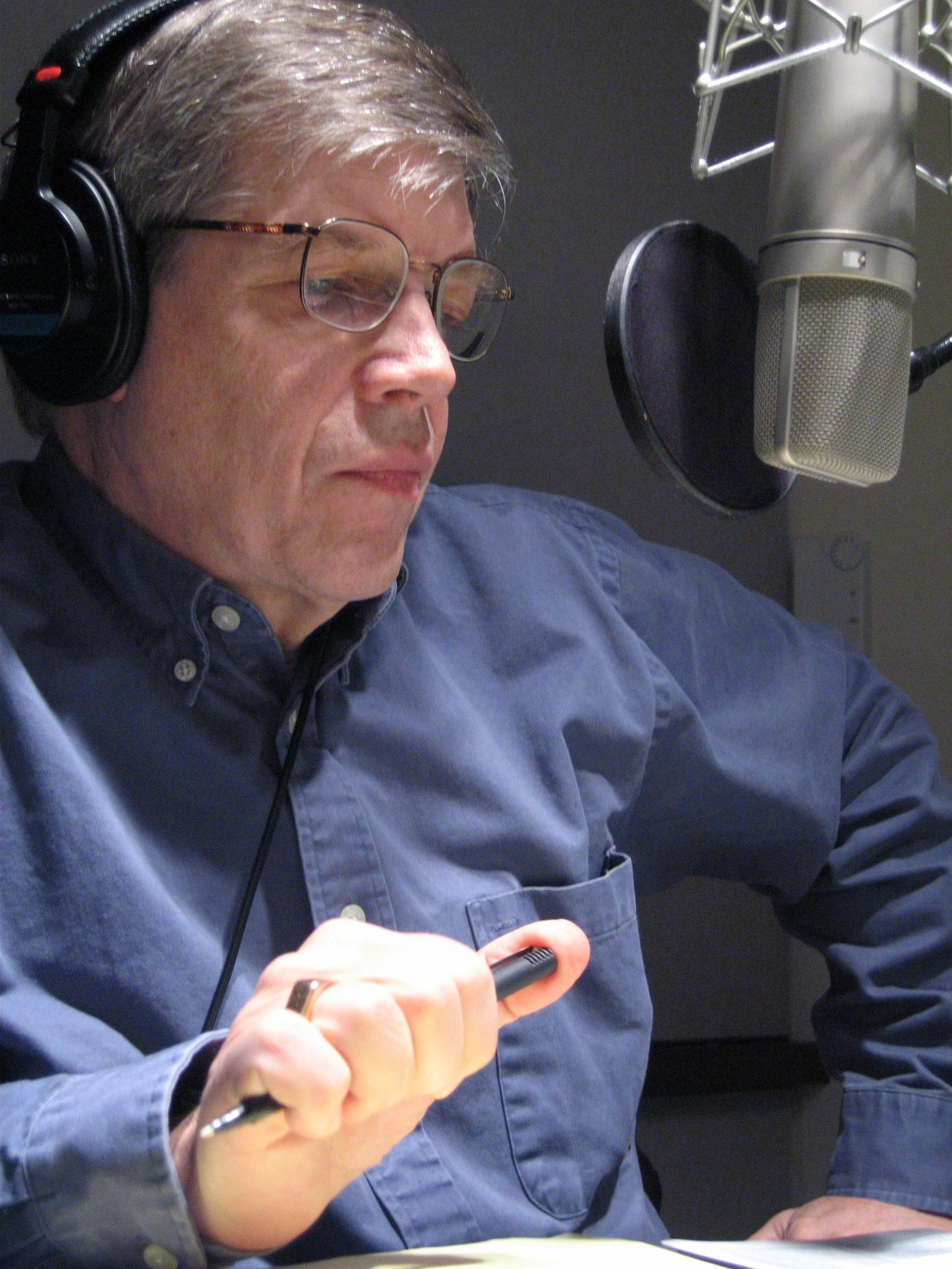 Rich Kleinfeldt at the microphone in a recording booth, headphones on, holding a script light.